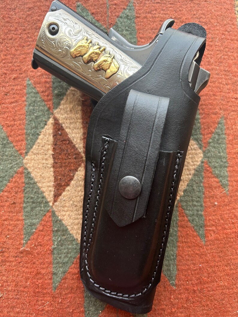 May include: A black leather holster with a stitched edge and a snap closure. A silver and gold handgun with an engraved design is inside the holster.