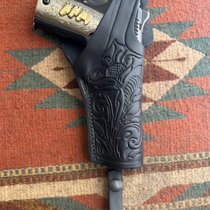 May include: Black leather holster with floral tooling, holding a silver and gold handgun. The handgun has gold lettering on the side. The holster is attached to a black leather strap.