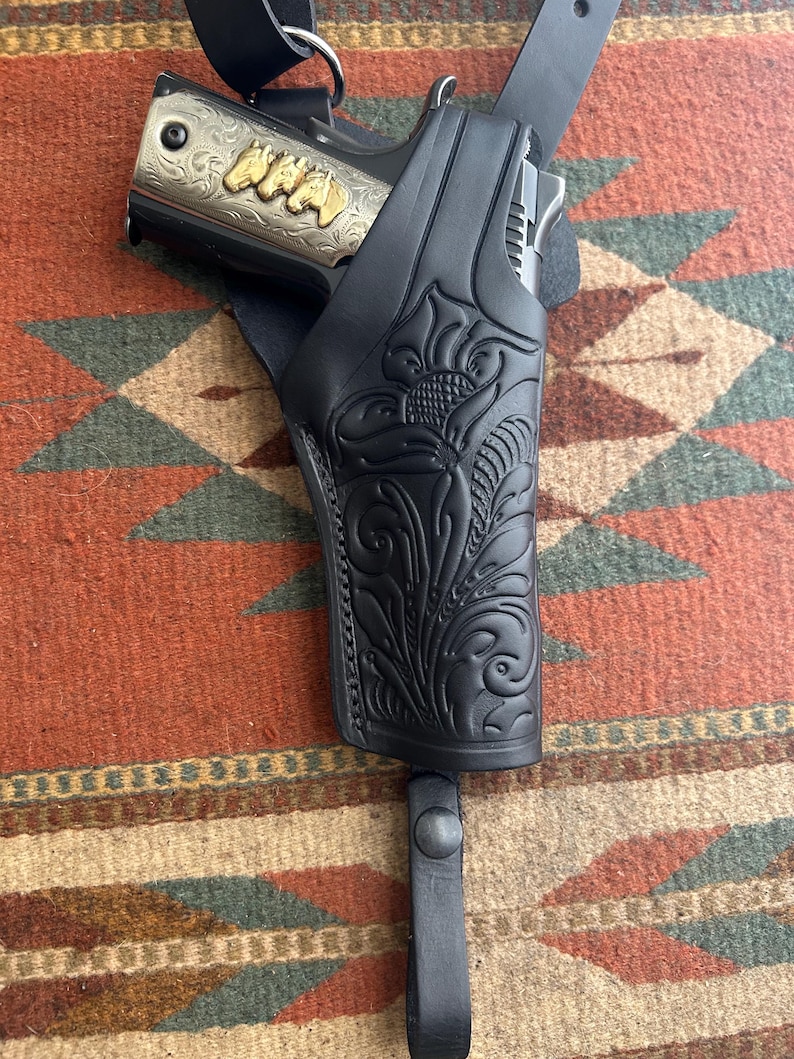 May include: Black leather holster with floral tooling, holding a silver and gold handgun. The handgun has gold lettering on the side. The holster is attached to a black leather strap.