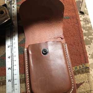 May include: A brown leather pouch with a white stitched border and a snap closure. The pouch is open and the inside is visible.