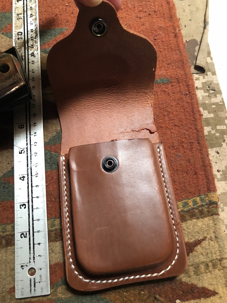 May include: A brown leather pouch with a white stitched border and a snap closure. The pouch is open and the inside is visible.