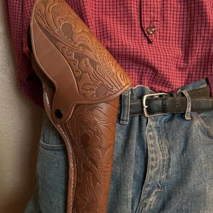 May include: A brown leather holster with floral tooling, worn on a person's hip. The holster is secured by a black button and white stitching. The person is wearing blue jeans, a black belt, and a red plaid shirt.