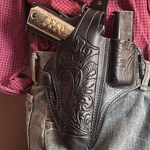 Puede incluir: Una funda de cuero negro con grabado floral y un portacargador a juego, sujeta al cinturón. La funda contiene una pistola semiautomática de tono plateado con incrustaciones doradas de cabezas de caballo en la corredera y grabados detallados.