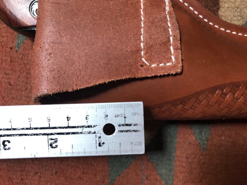 Op de afbeelding: Een bruine leren holster met witte stiksels. De holster wordt gemeten met een liniaal, wat een lengte van 7,6 cm laat zien.