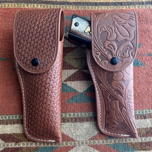 May include: Two brown leather holsters. One has a woven texture, the other features a floral design. Both have a black snap closure and white stitching. One holster contains a silver and gold handgun.