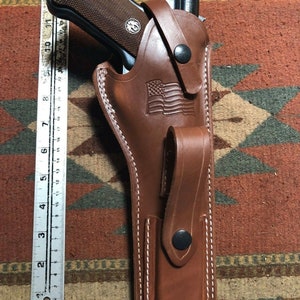 Puede incluir: Una funda de cuero marr&oacute;n con detalles de costura blanca. La funda tiene una solapa con un bot&oacute;n negro y un peque&ntilde;o bolsillo con un bot&oacute;n negro. La funda est&aacute; dise&ntilde;ada para sostener un arma de fuego. Una bandera estadounidense est&aacute; grabada en la funda.