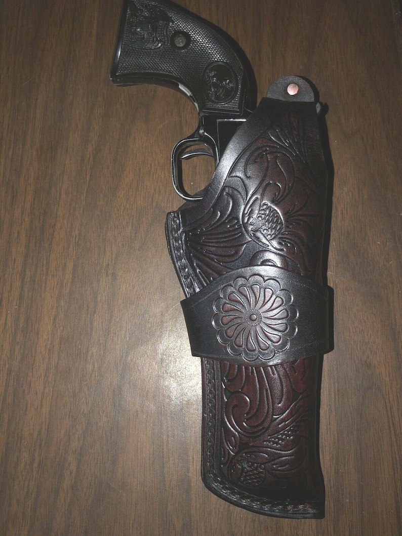 May include: A brown leather holster with intricate floral designs and a black revolver inside. The holster has a decorative metal buckle.