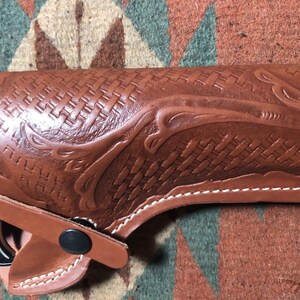 Op de afbeelding: Een bruine leren holster met een geweven patroon en een decoratief ontwerp. De holster heeft witte stiksels en een zwarte drukknoop sluiting.