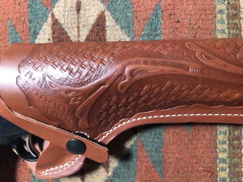 Op de afbeelding: Een bruine leren holster met een geweven patroon en een decoratief ontwerp. De holster heeft witte stiksels en een zwarte drukknoop sluiting.