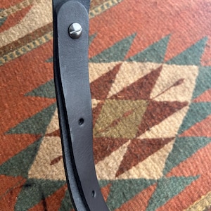 May include: Close-up of a black leather belt with a silver-colored screw and two holes. The belt is curved and appears to be made of high-quality leather. The background is a patterned rug with red, green, and tan colors.
