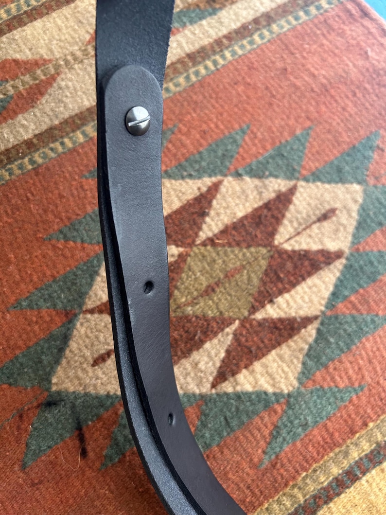 May include: Close-up of a black leather belt with a silver-colored screw and two holes. The belt is curved and appears to be made of high-quality leather. The background is a patterned rug with red, green, and tan colors.