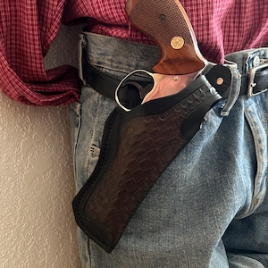 Op de afbeelding: Een bruine en zilveren revolver met een houten handvat is gedeeltelijk zichtbaar, in een zwart leren holster. Het holster heeft een textuurpatroon. De revolver is bevestigd aan een zwarte riem die met een spijkerbroek wordt gedragen.
