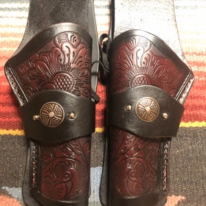 May include: A pair of brown leather gun holsters with intricate floral designs and silver metal accents. The holsters are attached to a brown leather belt.