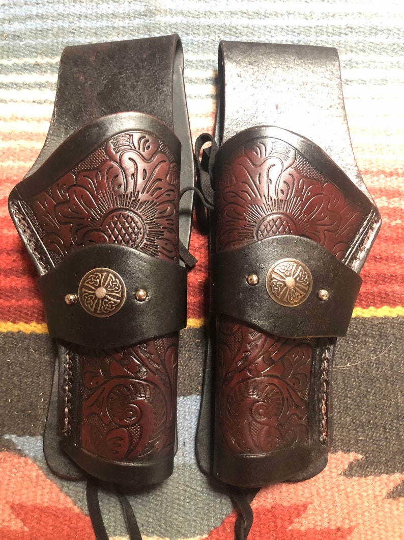 May include: A pair of brown leather gun holsters with intricate floral designs and silver metal accents. The holsters are attached to a brown leather belt.