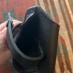 May include: A black leather holster with a loop and strap. The holster is designed to hold a firearm. The leather appears to be textured and the stitching is visible. The holster is shown against a patterned rug.