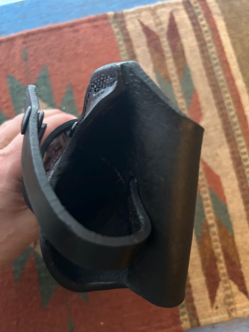 May include: A black leather holster with a loop and strap. The holster is designed to hold a firearm. The leather appears to be textured and the stitching is visible. The holster is shown against a patterned rug.