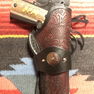 Puede incluir: Una funda de cuero marrón con intrincados diseños florales y una concha de plata. Una pistola plateada y negra está en la funda.