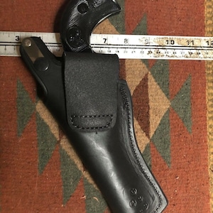 Pode incluir: Um coldre de couro preto para uma pistola. O coldre tem uma aba que cobre o guarda-mato e um passador de cinto.