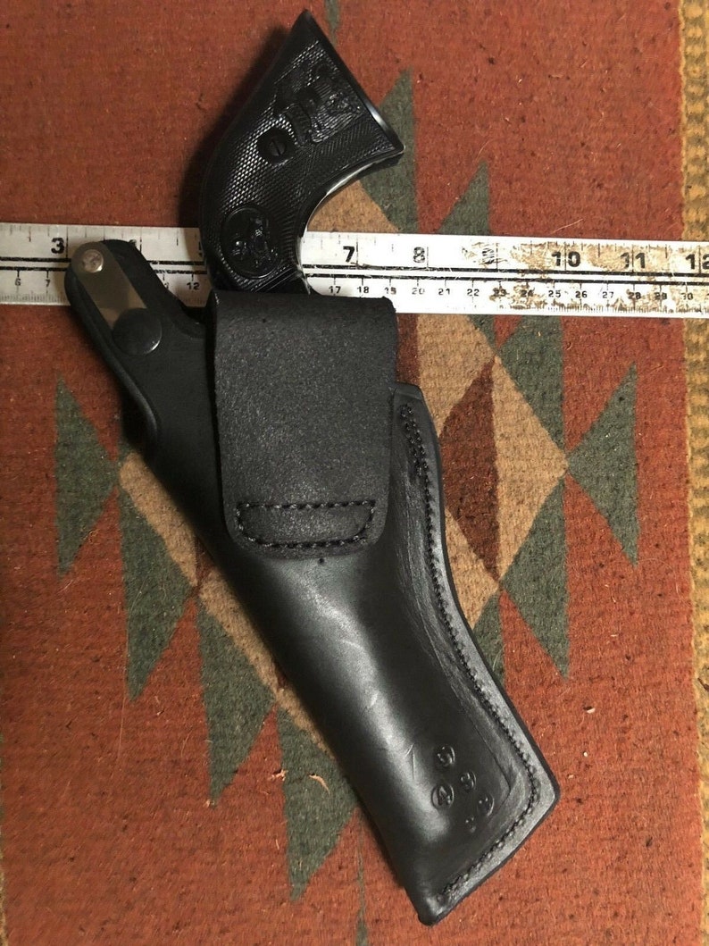 Pode incluir: Um coldre de couro preto para uma pistola. O coldre tem uma aba que cobre o guarda-mato e um passador de cinto.