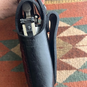 May include: A black leather holster containing a silver handgun. The holster features a belt loop and is held by a person. The leather is thick and textured.