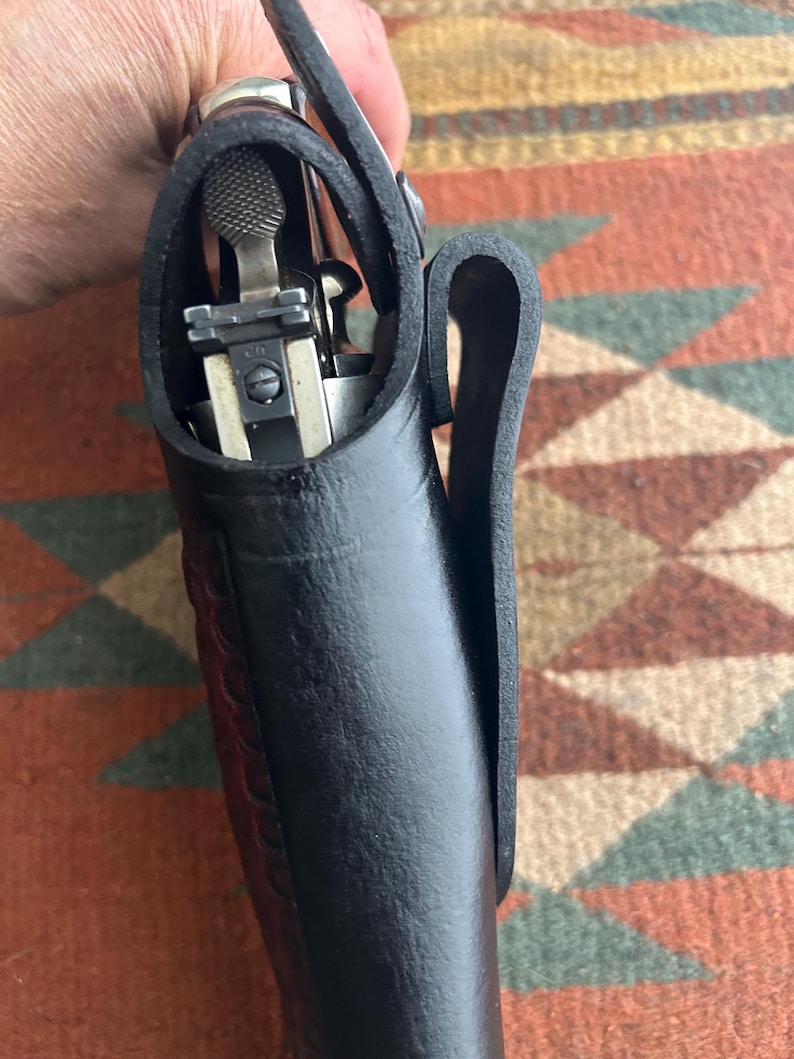 May include: A black leather holster containing a silver handgun. The holster features a belt loop and is held by a person. The leather is thick and textured.