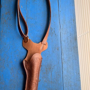 Puede incluir: Una funda de cuero marr&oacute;n con un dise&ntilde;o floral. La funda est&aacute; unida a una correa de cuero marr&oacute;n que se puede llevar sobre el hombro.