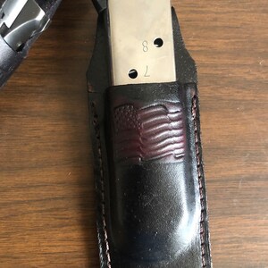 Puede incluir: Un portacargadores de cuero negro con un dise&ntilde;o de bandera americana de cuero marr&oacute;n. El portacargadores tiene un borde cosido y est&aacute; dise&ntilde;ado para sostener un cargador plateado con el n&uacute;mero "78" grabado en &eacute;l.