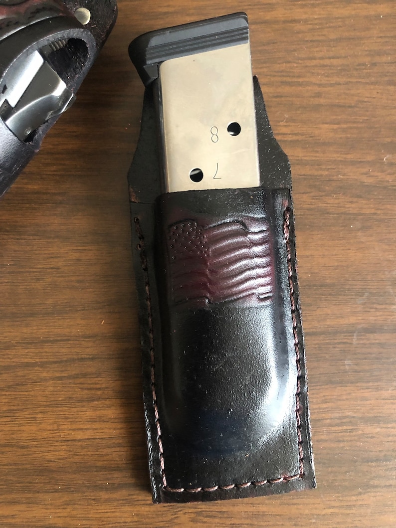 Puede incluir: Un portacargadores de cuero negro con un dise&ntilde;o de bandera americana de cuero marr&oacute;n. El portacargadores tiene un borde cosido y est&aacute; dise&ntilde;ado para sostener un cargador plateado con el n&uacute;mero "78" grabado en &eacute;l.
