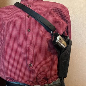 May include: Black leather shoulder holster with a decorative floral pattern. The holster is designed to hold a handgun. The strap is black and adjustable. The mannequin is wearing a red plaid shirt and blue jeans.