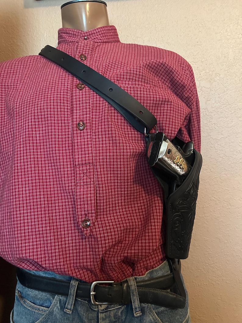 May include: Black leather shoulder holster with a decorative floral pattern. The holster is designed to hold a handgun. The strap is black and adjustable. The mannequin is wearing a red plaid shirt and blue jeans.