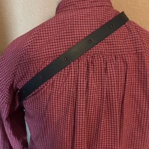 May include: A black leather strap with multiple holes is draped across the back of a red plaid shirt. The shirt is buttoned up and has a collar. The strap appears to be a shoulder strap for a bag or other accessory.