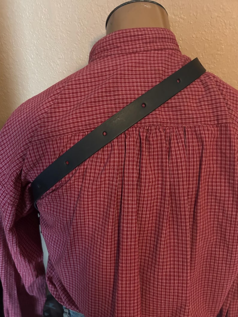 May include: A black leather strap with multiple holes is draped across the back of a red plaid shirt. The shirt is buttoned up and has a collar. The strap appears to be a shoulder strap for a bag or other accessory.