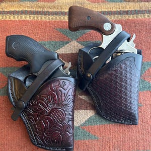 May include: Two revolvers in black leather holsters. One holster features a floral design, the other a basketweave pattern. The revolvers have brown grips and are set against a patterned rug.