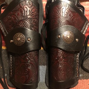 May include: Two brown leather holsters with intricate floral designs and metal accents. The holsters are designed to hold pistols and have a strap for securing them to a belt.