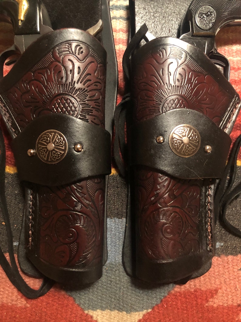 May include: Two brown leather holsters with intricate floral designs and metal accents. The holsters are designed to hold pistols and have a strap for securing them to a belt.