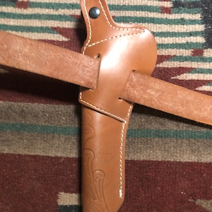 May include: A brown leather holster with a decorative design and a snap closure. The holster is attached to a brown leather belt.