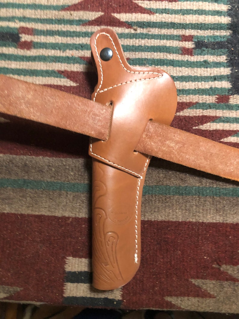 May include: A brown leather holster with a decorative design and a snap closure. The holster is attached to a brown leather belt.