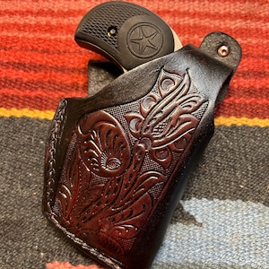May include: A brown leather holster with a floral design, holding a small silver handgun with a black grip.
