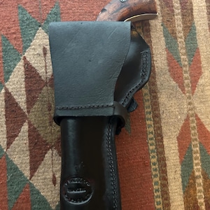 May include: Black leather holster with a stitched design, holding a vintage pistol with a wooden handle. The holster has a stamped logo and is secured with a strap. The pistol's barrel is visible.