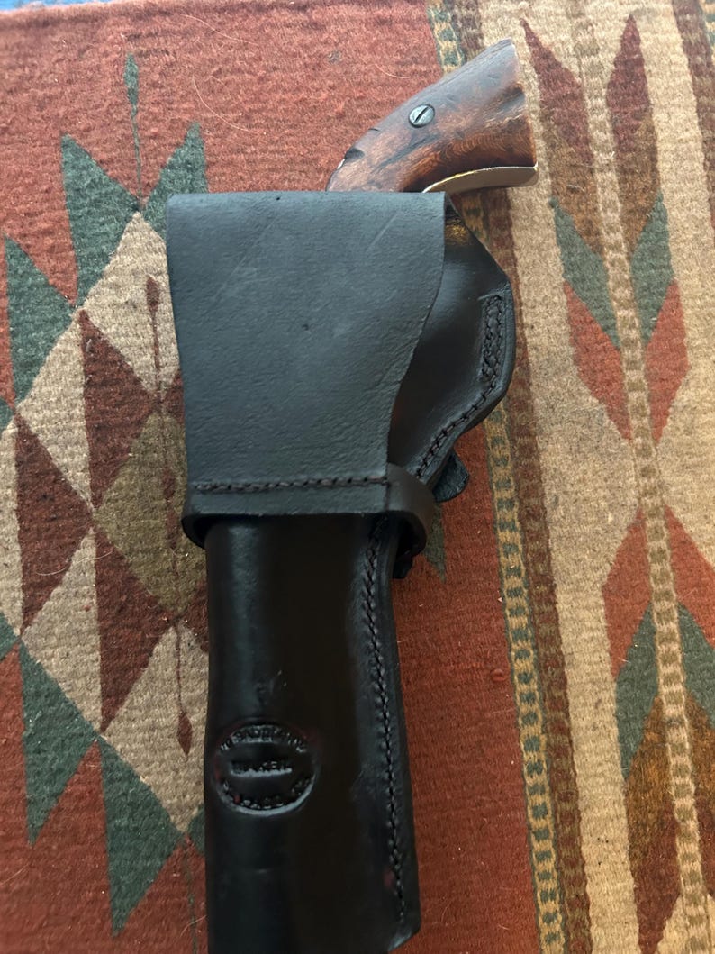 May include: Black leather holster with a stitched design, holding a vintage pistol with a wooden handle. The holster has a stamped logo and is secured with a strap. The pistol's barrel is visible.