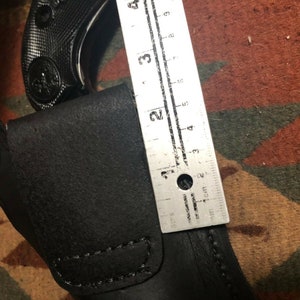 Pode incluir: Um coldre de couro preto com fecho de press&atilde;o prateado. Uma arma de fogo preta &eacute; parcialmente vis&iacute;vel no coldre. Uma r&eacute;gua &eacute; vis&iacute;vel &agrave; direita do coldre, medindo 5 cm.