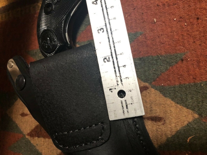 Pode incluir: Um coldre de couro preto com fecho de press&atilde;o prateado. Uma arma de fogo preta &eacute; parcialmente vis&iacute;vel no coldre. Uma r&eacute;gua &eacute; vis&iacute;vel &agrave; direita do coldre, medindo 5 cm.