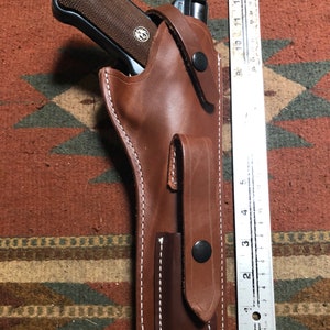 May include: A brown leather holster with a white stitch detail and a black snap button. A silver handgun is inside the holster.