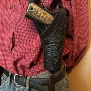 May include: Black leather shoulder holster with floral tooling, holding a silver handgun with gold horse head accents. The holster is worn over a red plaid shirt and blue jeans, secured by a black belt.