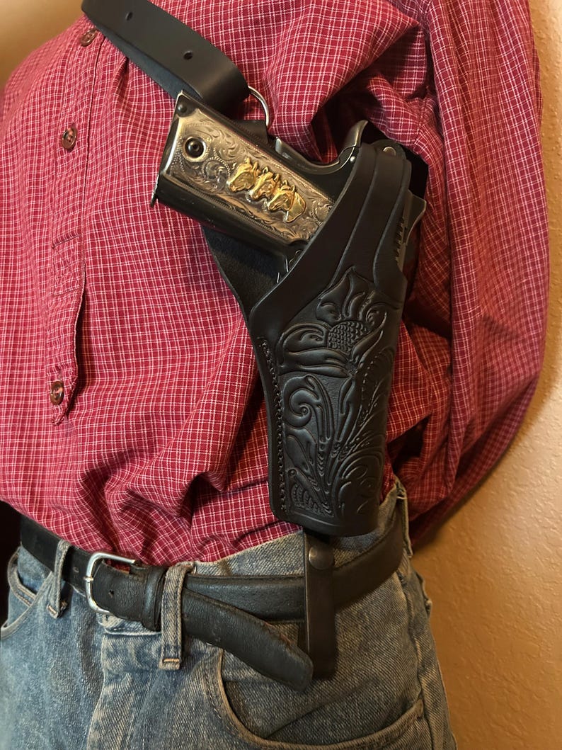May include: Black leather shoulder holster with floral tooling, holding a silver handgun with gold horse head accents. The holster is worn over a red plaid shirt and blue jeans, secured by a black belt.