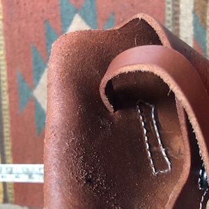 Op de afbeelding: Een bruine leren holster met witte stiksels en een zwarte knop. De holster heeft een band die kan worden gebruikt om hem aan een riem te bevestigen.