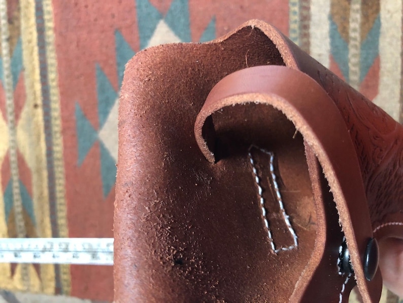 Op de afbeelding: Een bruine leren holster met witte stiksels en een zwarte knop. De holster heeft een band die kan worden gebruikt om hem aan een riem te bevestigen.