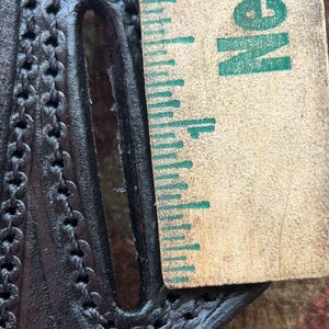 K&ouml;nnte beinhalten: Nahaufnahme eines schwarzen Lederholsters mit einem gestickten Design. Ein Lineal mit dem aufgedruckten Wort "NEW" liegt neben dem Holster.