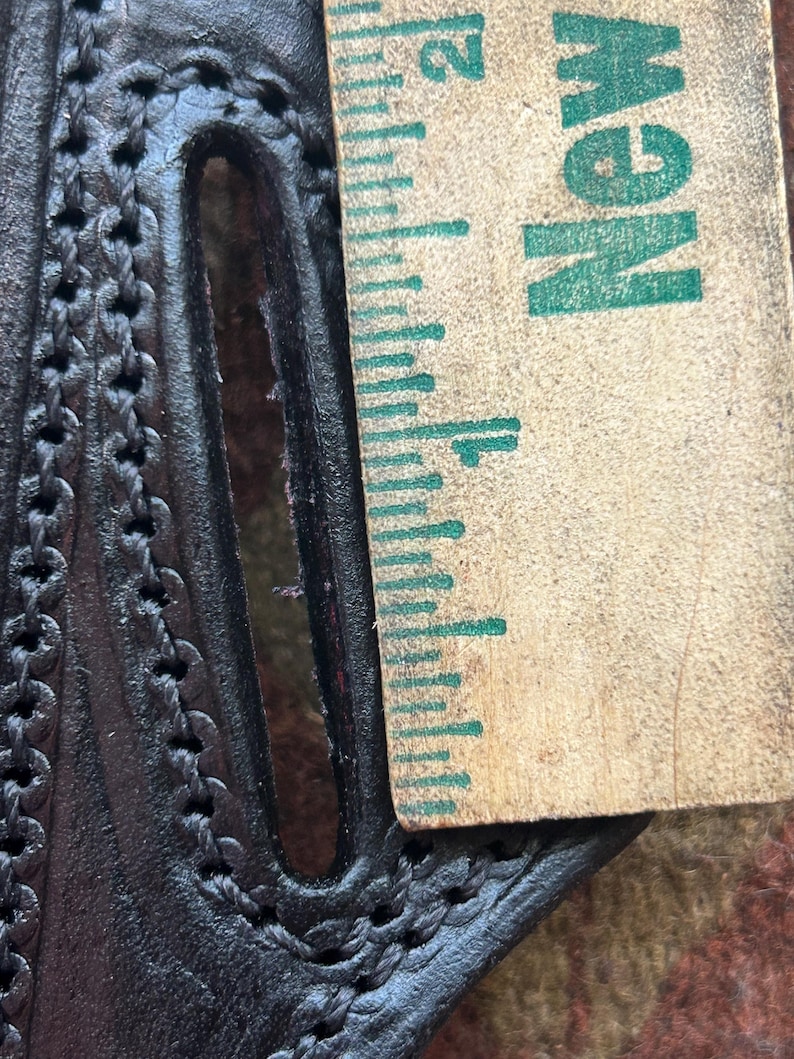 K&ouml;nnte beinhalten: Nahaufnahme eines schwarzen Lederholsters mit einem gestickten Design. Ein Lineal mit dem aufgedruckten Wort "NEW" liegt neben dem Holster.