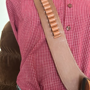 Pode incluir: Um coldre de pistola de couro marrom com um la&ccedil;o de muni&ccedil;&atilde;o de couro marrom. O coldre &eacute; usado sobre uma camisa xadrez vermelha e branca.
